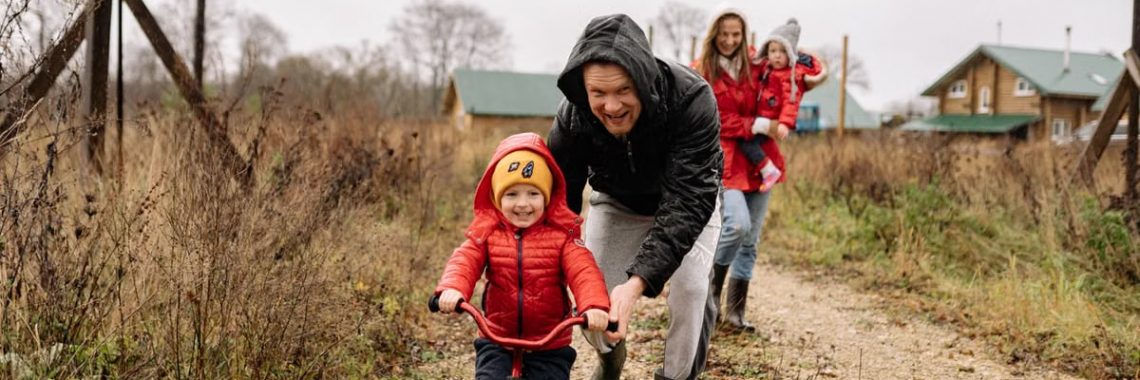 A happy family enjoys a bonding moment outdoors with a child learning to ride a bicycle on a fall day.